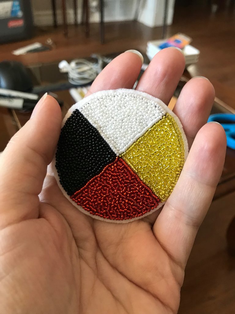 Beaded Medicine Wheel in Cynthia's left hand. The top or northern quadrant is white, the right side or eastern quadrant is yellow, the bottom or south quadrant is red, and the left side or western quadrant is black.