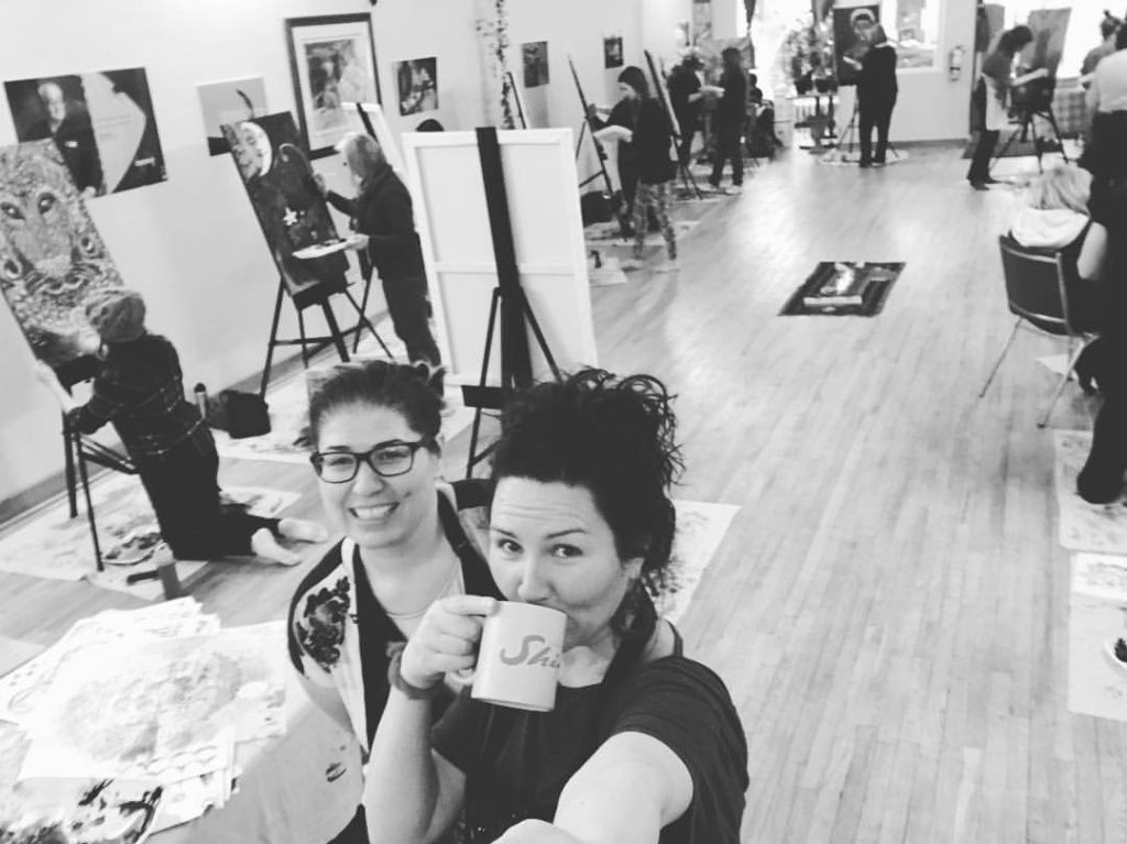 Angie is in the foreground, having a sip from a mug. Cynthia is immediately behind her. They're both looking at the camera. The class of students at their easels are in the background. Photo from March 2017 at Blue Sky Community Healing Centre.