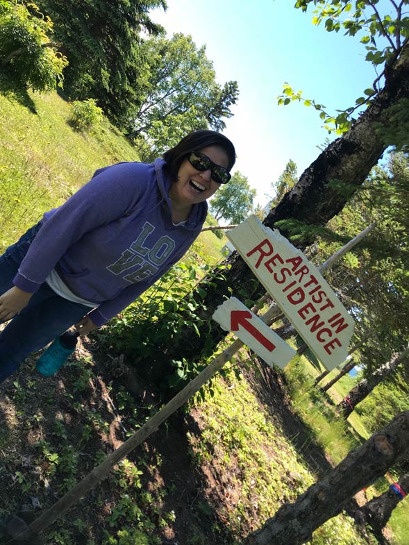 diagonally taken photo showing Cynthia standing on the grass, next to a large tree trunk and a sign that says "Artist in Residence", with an arrow, pointing at Cynthia. She's wearing big black sunglasses and a purple sweatshirt that has the four letters that spell love in a square pattern (L.O. on top and V.E. on the bottom). There are more trees further in the background and the sky is blue.